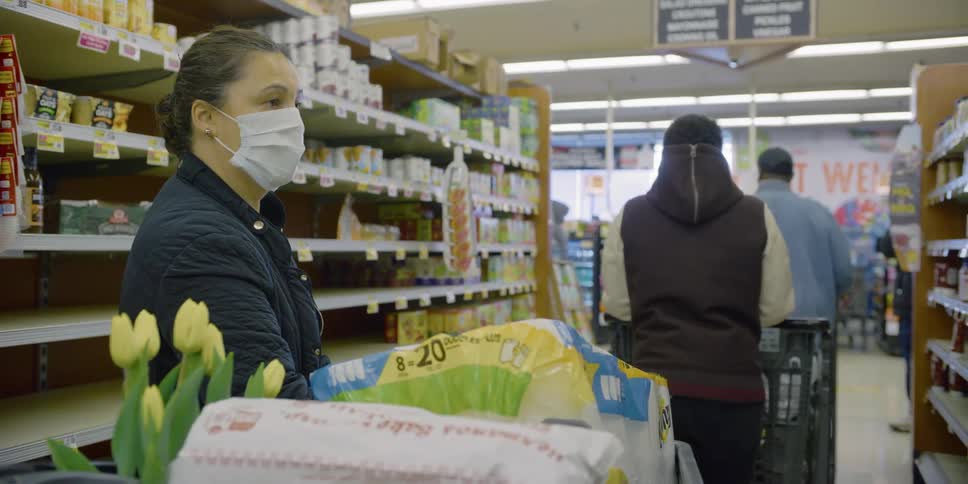A woman wearing a mask waits behind others in the cashier line which extends into the aisle due to social distancing
