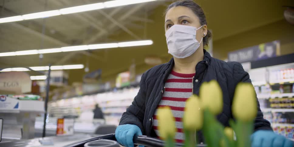 Woman shops through grocery store pushing cart and wearing gloves and a mask