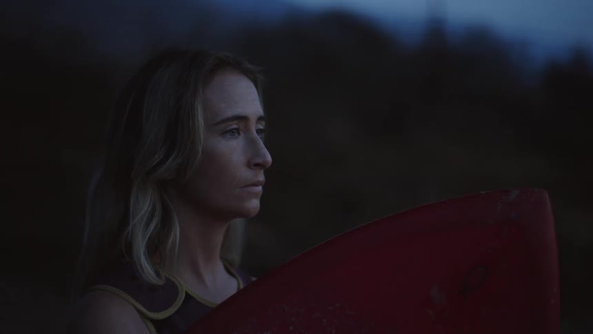 Blonde surfer stands with red surfboard near woods while looking out
