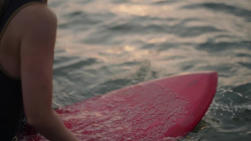 Woman in black wetsuit sits on red surfboard on water at sunset