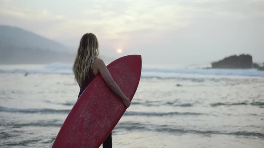 Blonde female surfer carries red surfboard out to ocean as fog obscures sun sitting near horizon