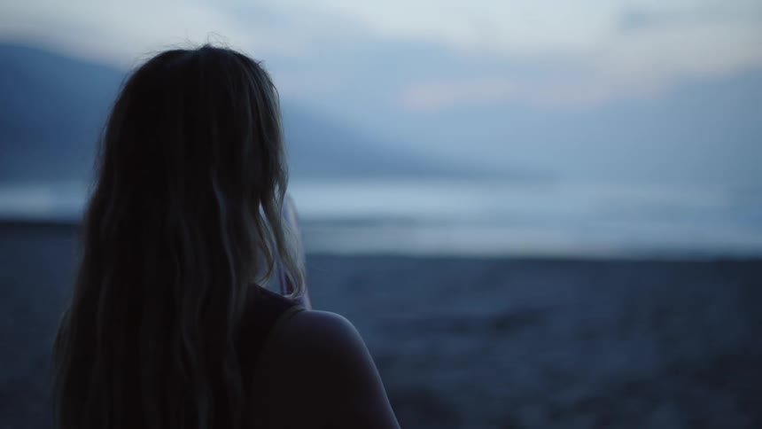 Young female surfer holding a surfboard looks at the ocean from the beach at evening