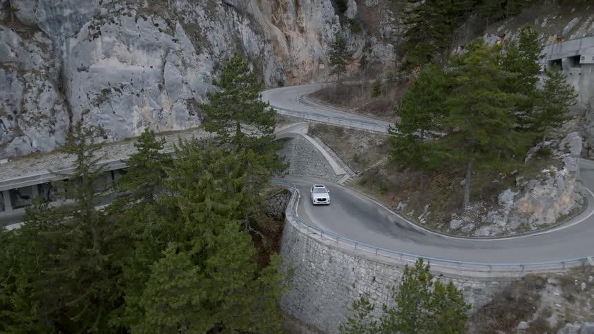 Aerial view tracking white car traveling along mountain pass and ascending mountain around hairpin turns and underpasses