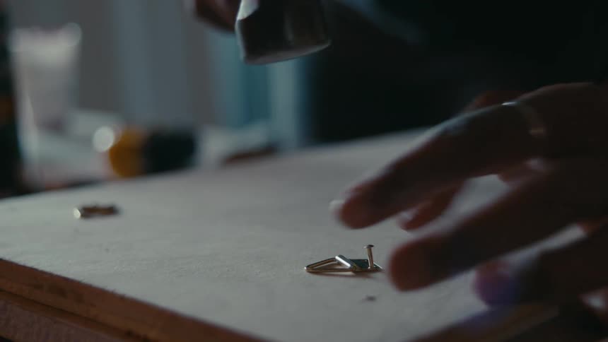 A persons hands are seen gently hammering a small nail into the back of picture to hold a hanger