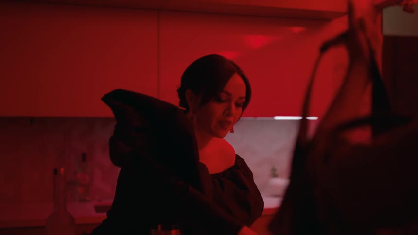 A woman carrying a drink walking into her kitchen as her friend follows her and sits down