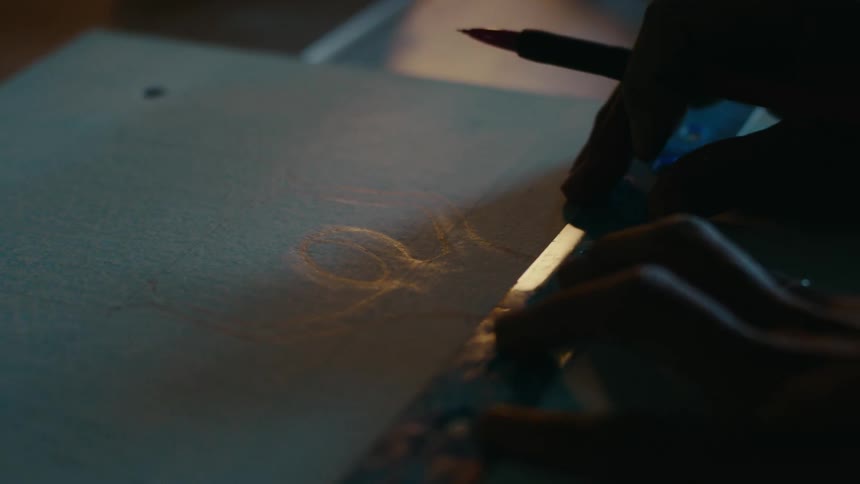 An artist sketches a humanshaped outline using a pencil and ruler on paper at a desk in a dark room
