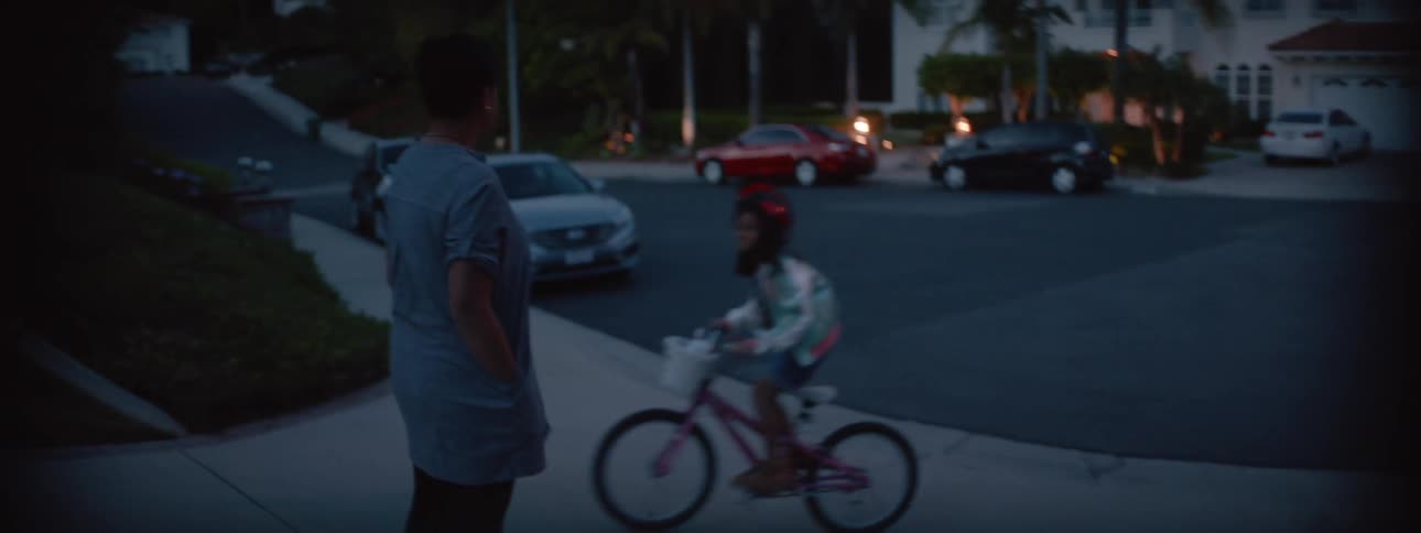 A mother stands watching her daughter ride her bicycle into their driveway