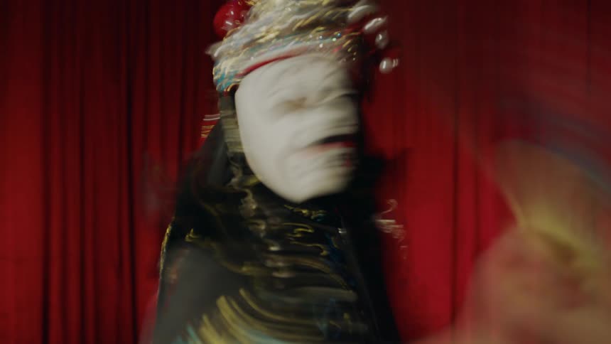 A person dressed in a costume with a mask and hat on dancing around a stage waving a fan