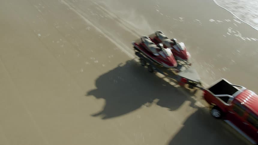 Red crewcab pickup truck towing two jet skiswhile driving along the beach directly on the sand and in the water
