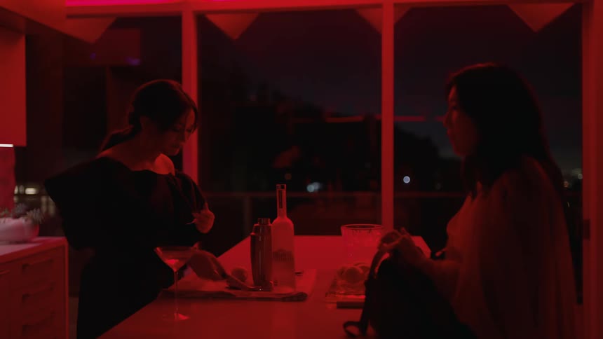 Two women talking over a counter in the kitchen while one prepares a drink with the kitchen lit up in red light