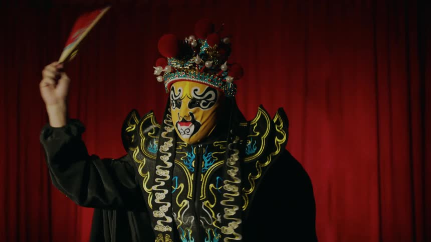A dancer on stage wearing a costume mask and hat while twirling around a fan