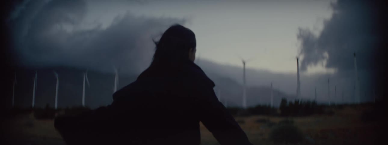 A woman is having difficulty walking against strong wind toward a wind farm as dark storm clouds move overhead