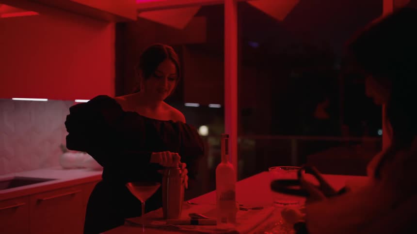 Friends talk in kitchen lit by colored lighting at night while one of them mixes and pours a cocktail