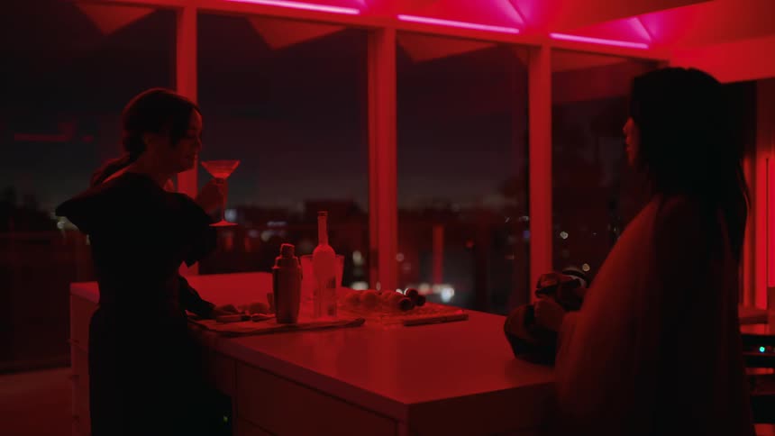 Two women standing in a kitchen one drinking and talking while the room is lit with red dim light