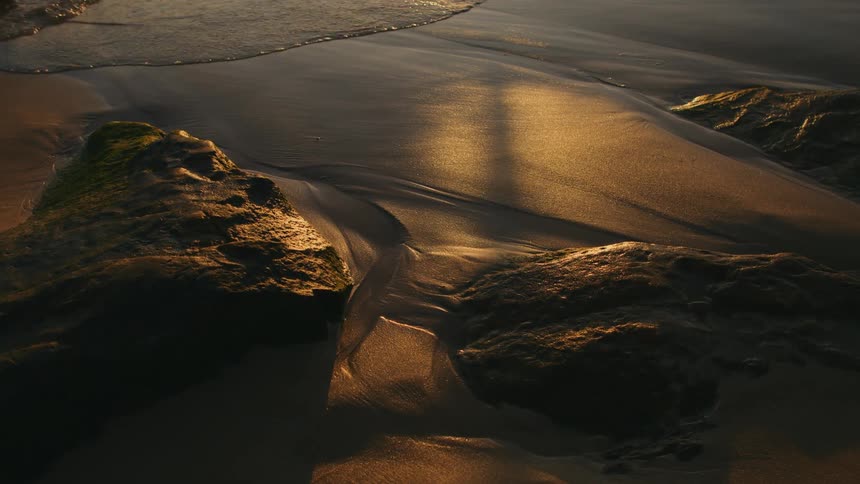 A shadow moves across the sand in the light next to large rocks as water creeps up from the edge of the sand than recedes