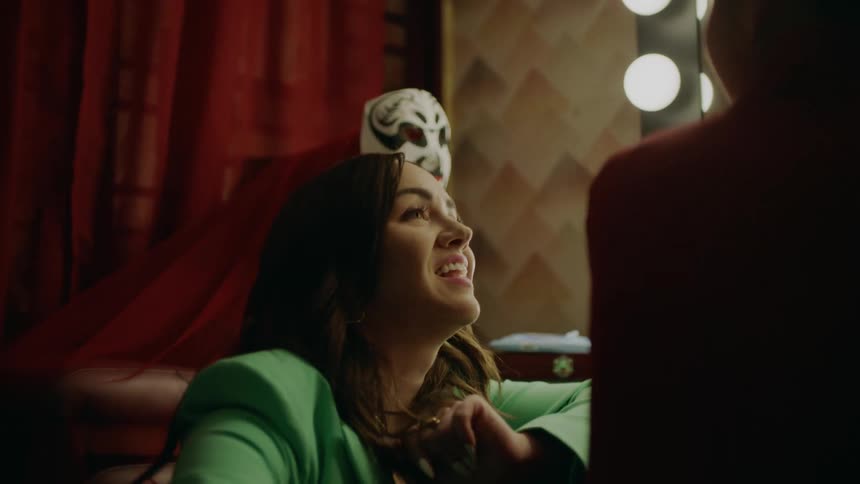 In a dressing room a woman kneels down and talks to another woman who is sitting at a vanity with a large mirror