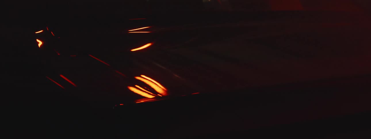 An up close of a shiny car surface and the back head light as light and shadows are cast across the car