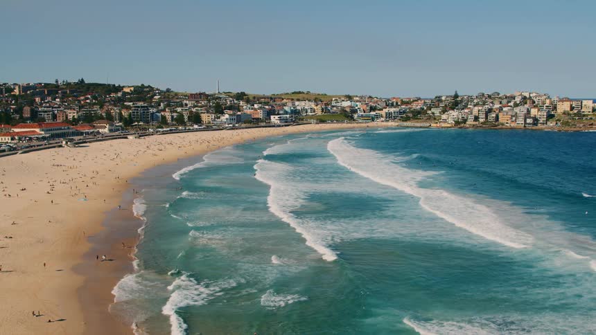 An aerial of a sandy beach with people on it and blue ocean waves rolling onto shore with suburban areas around the cove