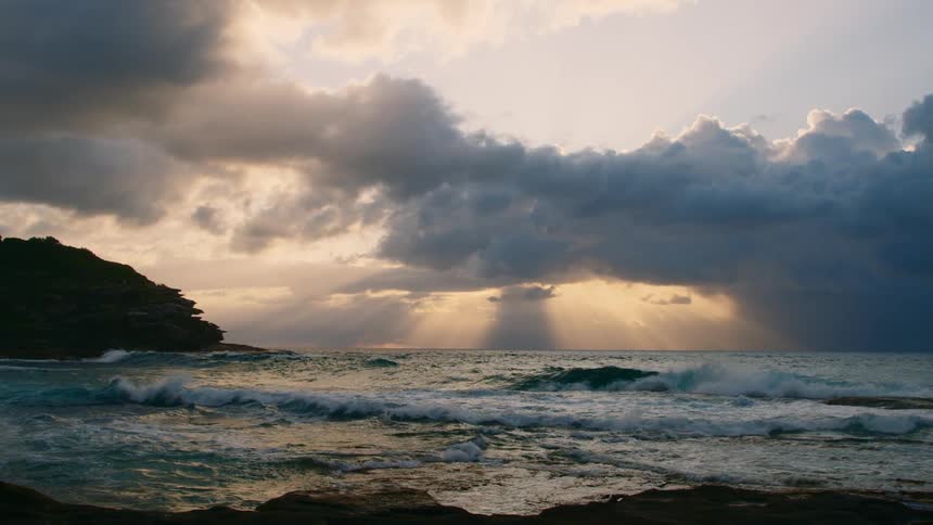 Sun rays casting through the clouds above the rough seas as waves crash against a coastline