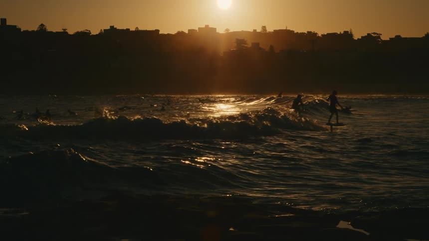 A group of surfers are swimming and riding the waves at sunset with a distant city in the background