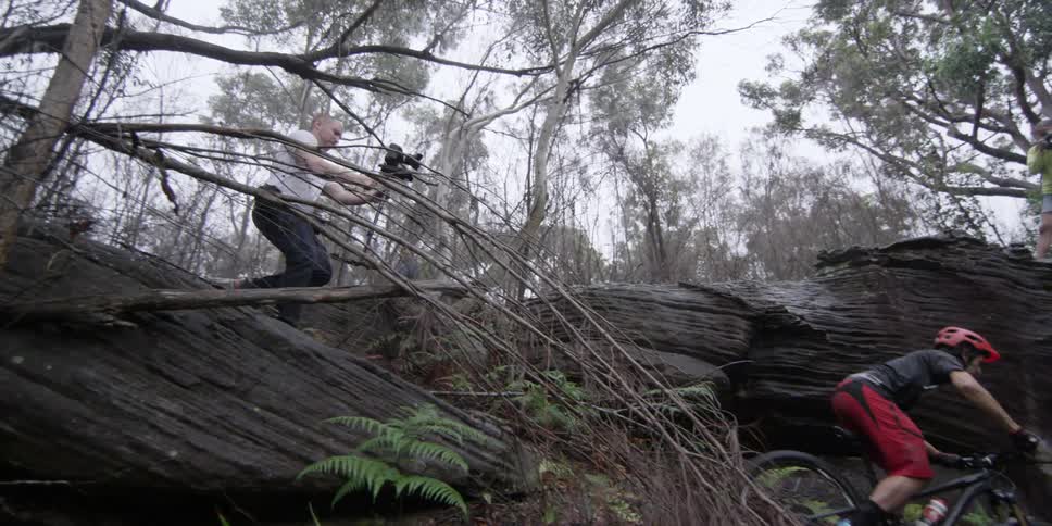 A cameraman filming a man riding a mountain bike along rough mountain forest terrain