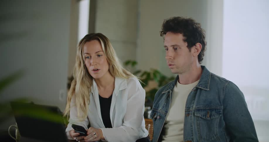 A man and a woman are sitting behind a laptop computer talking as she holds her phone in her hand