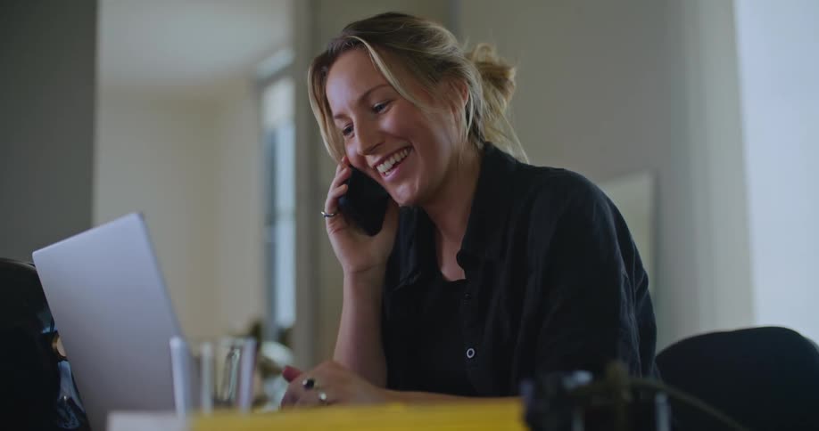 A woman is sitting at a desk with a laptop computer as she talks on the phone and waves her hand and smiles as she speaks