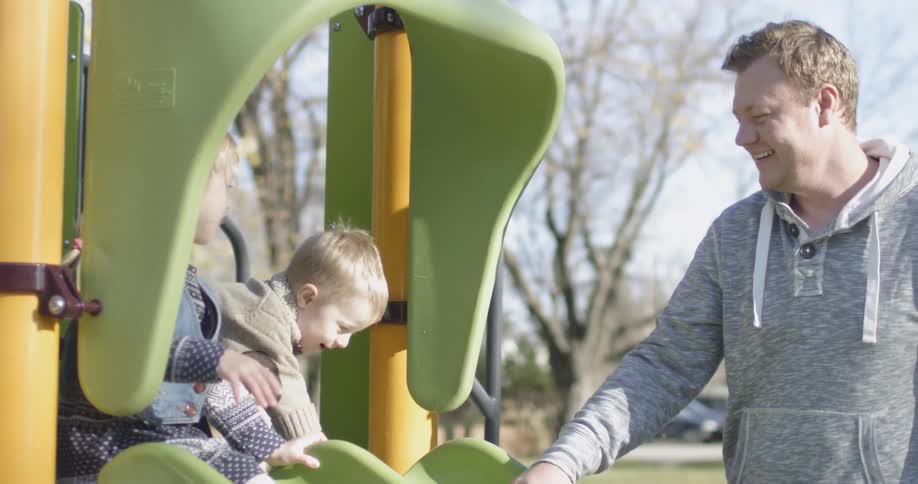 Children sit on the top of a slide at the park with their father smiling