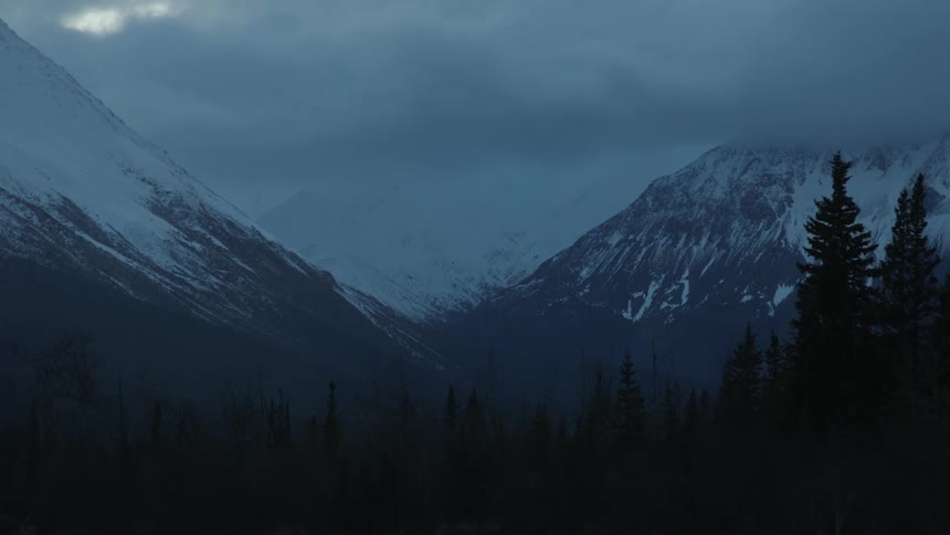 A forest of trees sit at the foot of snow covered mountains under a darkening cloudy sky