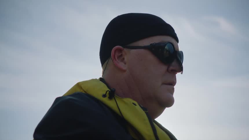 A man wearing a hat and sunglasses with a winter coat is standing outside as the sun shines through the clouds