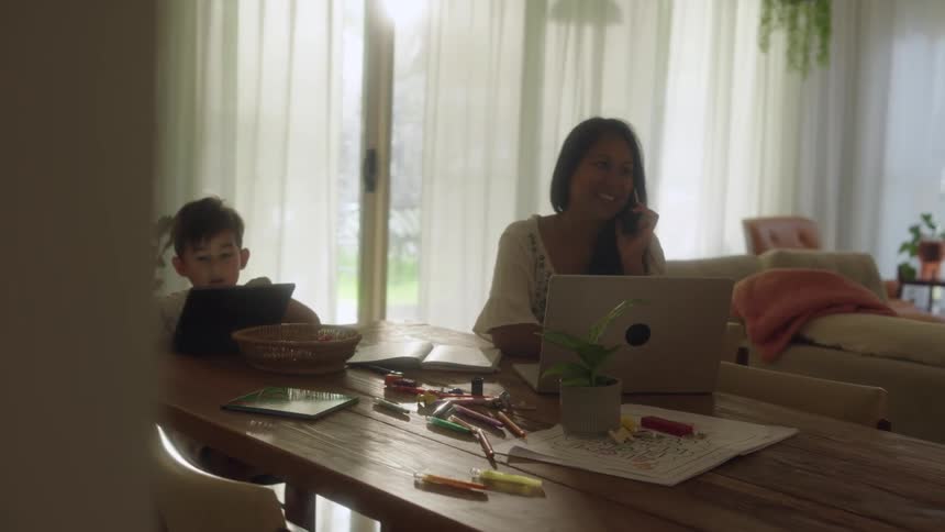 Mother on a phone call while using a laptop sitting next to her son on the dining room