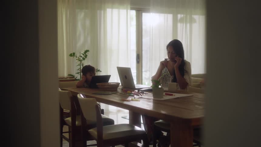 A mother and her son are sitting at a long table while she talks on her phone and looks at a laptop