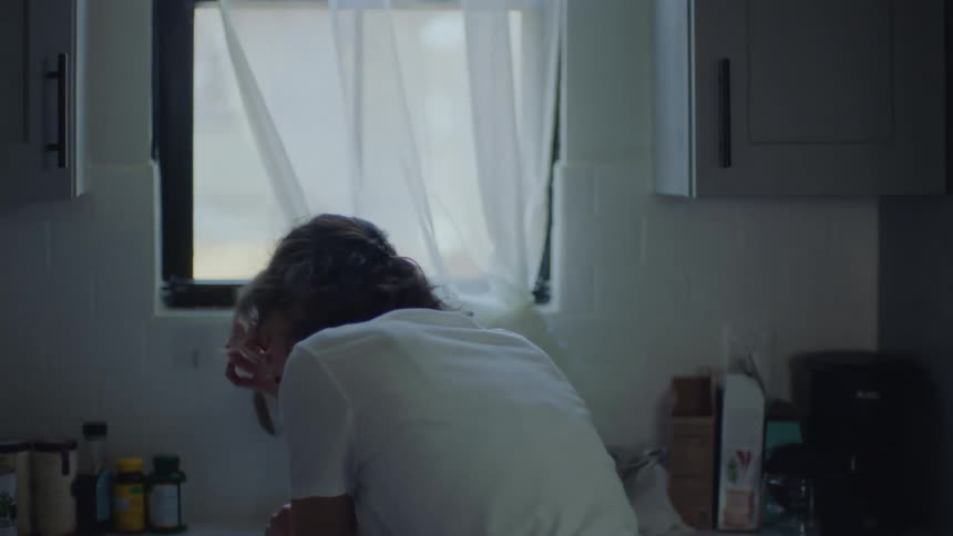 A woman standing in her kitchen preparing food then stopping and putting her elbows on the counter and her head in her hands