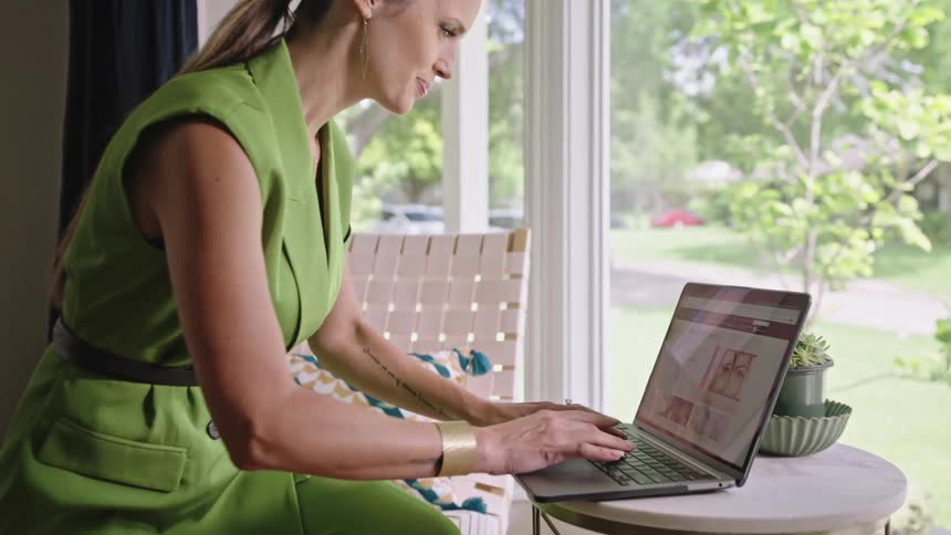 Woman wearing a green outfit grabs a laptop from a small table near a balcony