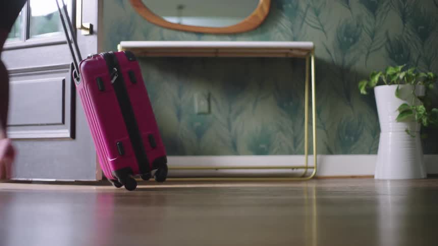 A woman walks through a room while pulling a pink suitcase and opens a door to go outside