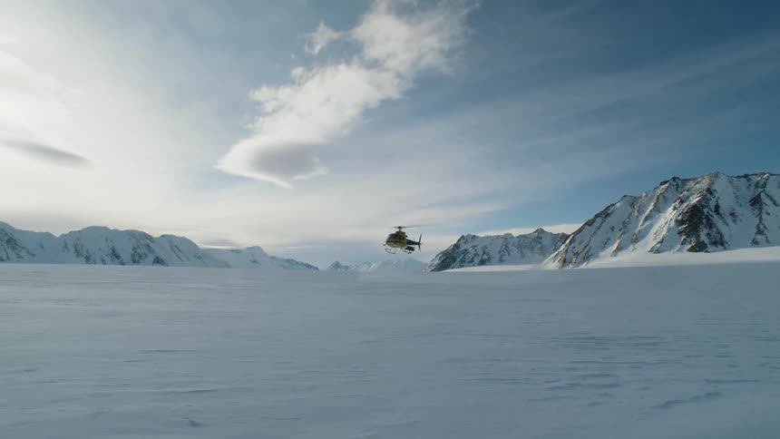 A helicopter flies between rocky cliffs to land on a snowy plain in an arctic environment