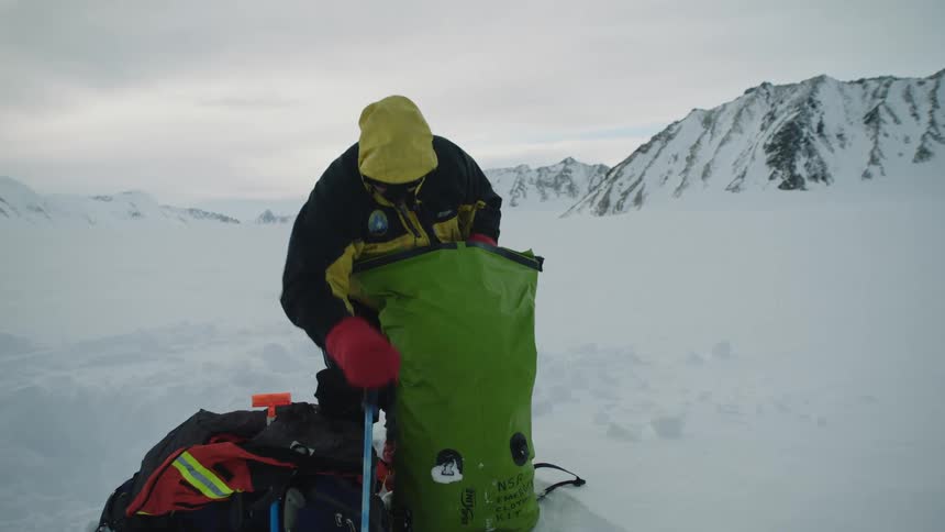Snow covers the ground and mountain cliffs behind a man in winter survival gear who is pulling climbing equipment out from his outdoor travel bag