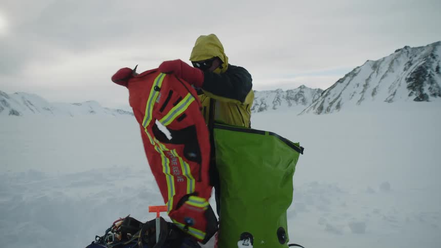 A man taking items out of a large green bag in the snow among mountain tops
