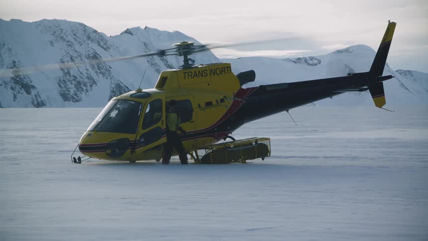 A helicopter is sitting on snow covered ground near mountains as the rotor spins a man opens the door and gets out