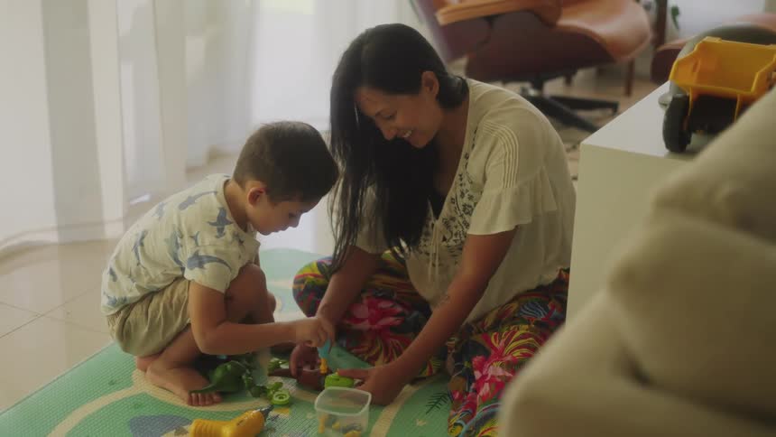Mother holds toy while son uses screwdriver on it as they play on floor in living room