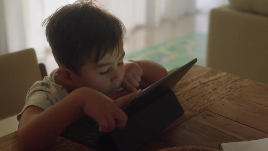 A little boy is sitting at a table with his hand on a tablet computer and his face is close to the screen