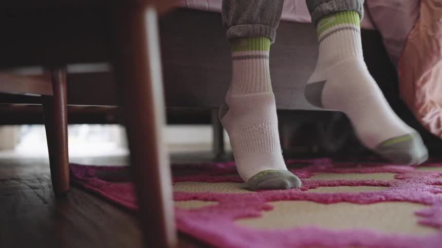 It is daylight and a persons feet appear at the side of bed both hitting the floor at the same time then lifting up and twisting at the ankle one at a time before the person with socks and sweatpants on stands up and walks away