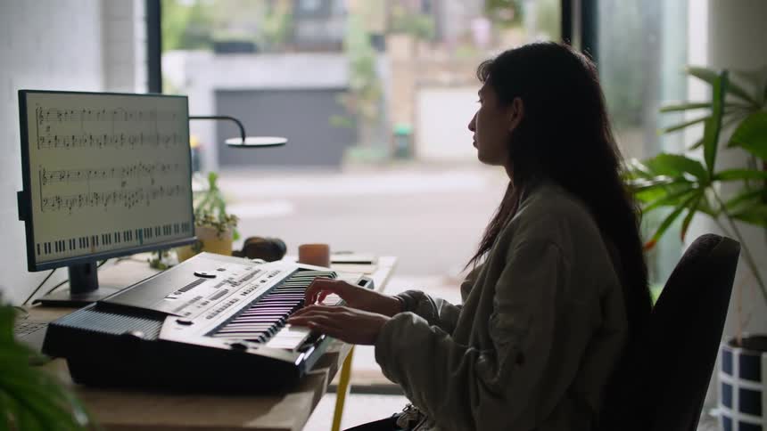 A teenage girl is sitting at a table near a window playing a keyboard in front of a computer monitor