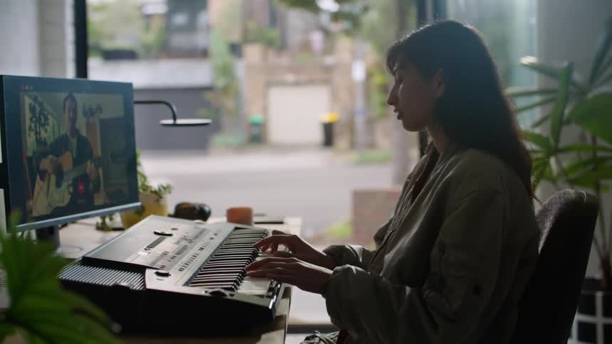 A woman sitting playing a piano keyboard while on video chat with a man playing the guitar