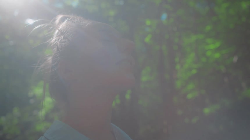 The sun shines upon a young woman standing outside near the woods with a smile on her face as she smells the fresh air