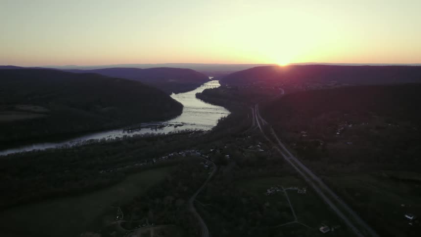 Aerial view of farming area with few houses beside a large river with a long highway beside it while the sun begins to set below the mountains in the distance