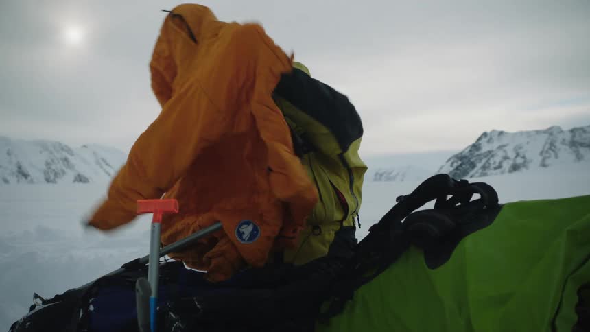 A man in a snow covered landscape is outdoors hurriedly unpacking an additional snow coat in the daytime