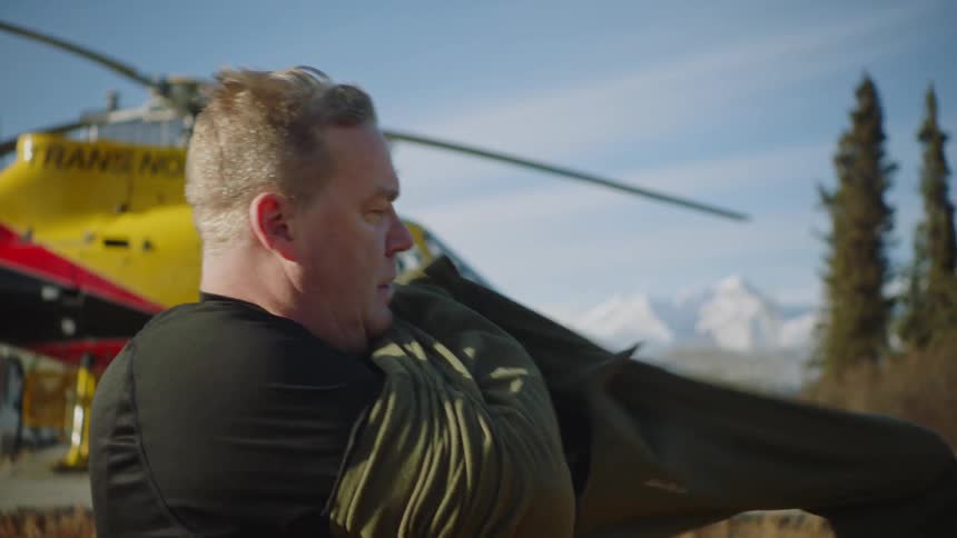 A man stands in front of a helicopter and puts on a pullover