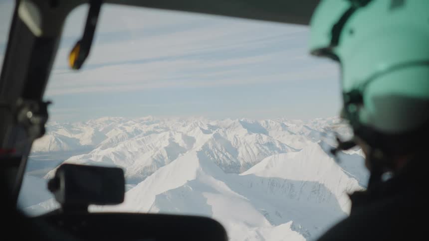 Pilot wearing a white helmet flies a helicopter over mountains covered in snow