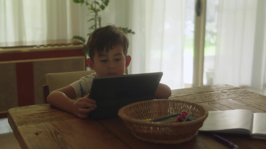 Boy with white shirt sits at table using tablet in the living room at home